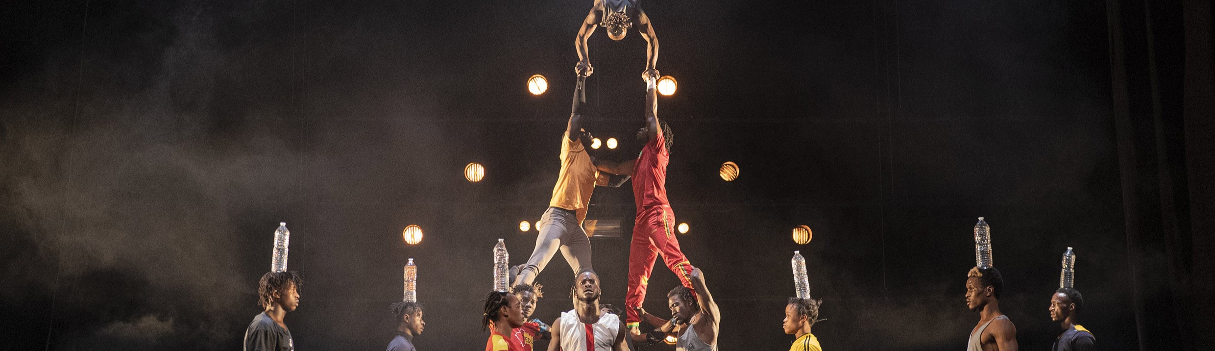 Grupp av människor utöver grupp akrobatik samtidigt som det står personer runt och balanserar vattenflaskor på huvudet.